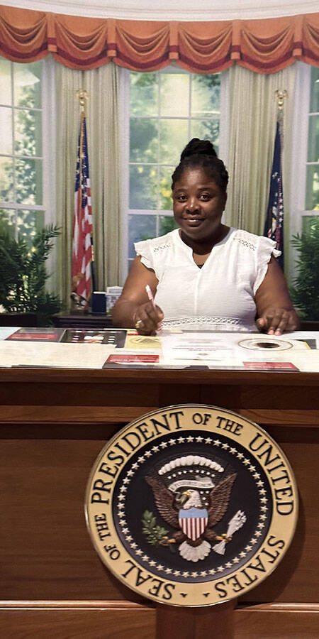 woman in a white dress seated at a replica of the President of the United States’ desk.