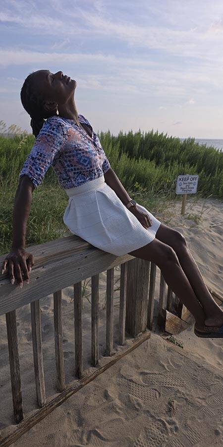 Black woman sitting on a fence on a sandy beach at sunset.