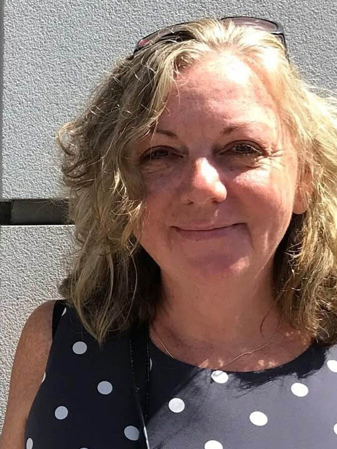 White woman making a tight-lipped smile,standing in gront of a grey wall