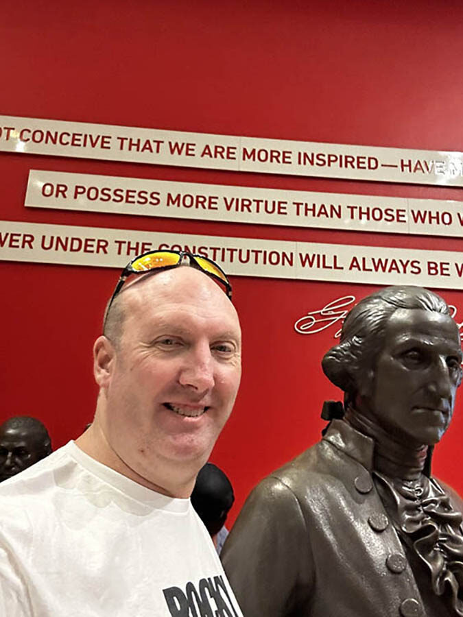 Smiling white man with sunglasses next to a bronze statue of one of the Founding Fathers.