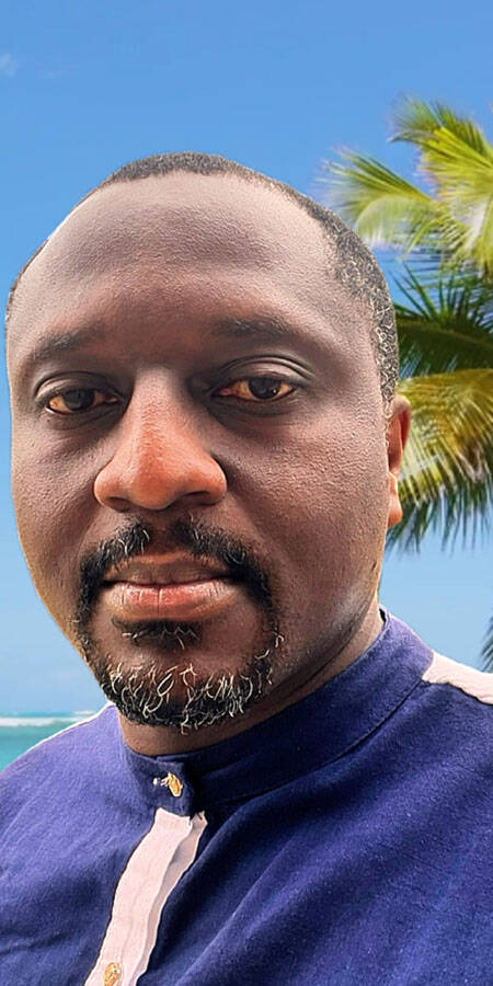 Black man in a collared shirt. A palm tree is visible in the background against a blue sky.