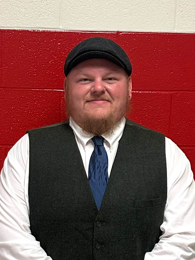 White man in a vest and tie, standingagainst a red-and white brick wall