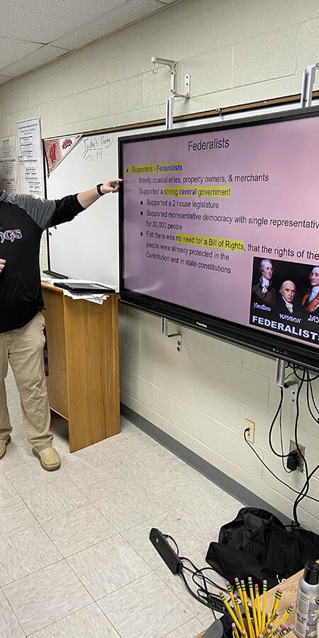 Teacher guesturing at a screen with a pink background.