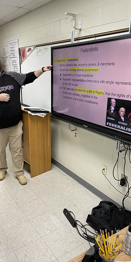 Teacher guesturing at a screen with a pink background.
