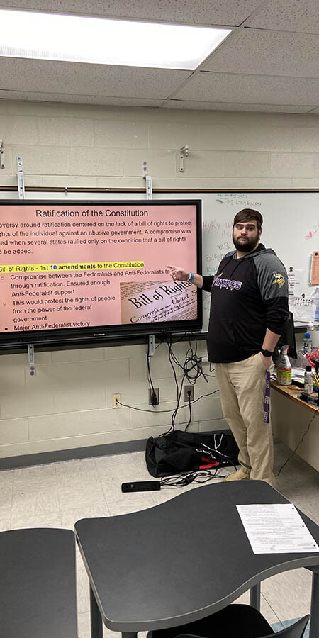 Teacher guesturing at a screen with a pink background.