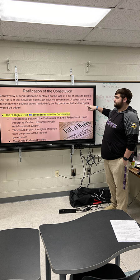 Teacher guesturing at a screen with a pink background.