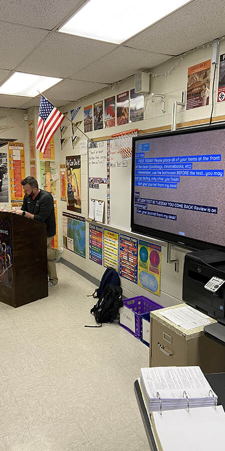 Teacher standing at a podium. A screen to the right shows highlighted text on a sepia background.