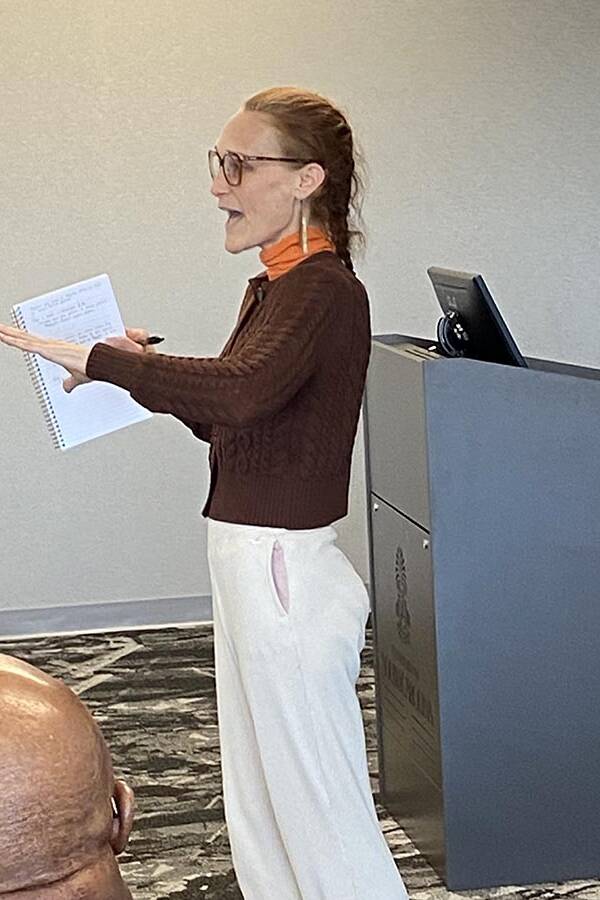 Woman in a brown sweater holding a folder in one hand an pointing with the other.