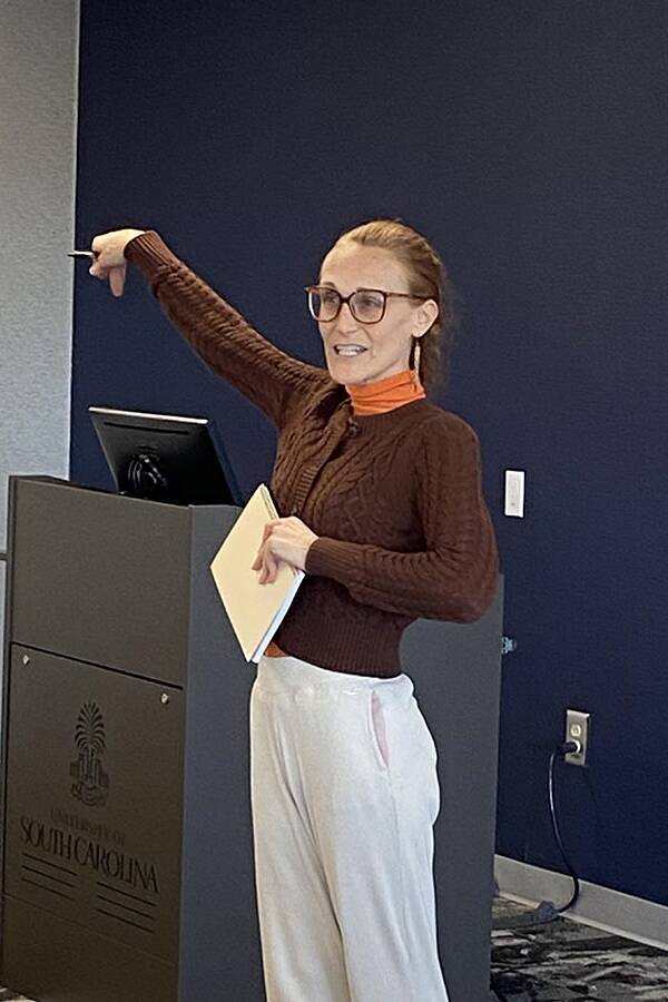 Woman in a brown sweater holding a folder in one hand an pointing with the other.