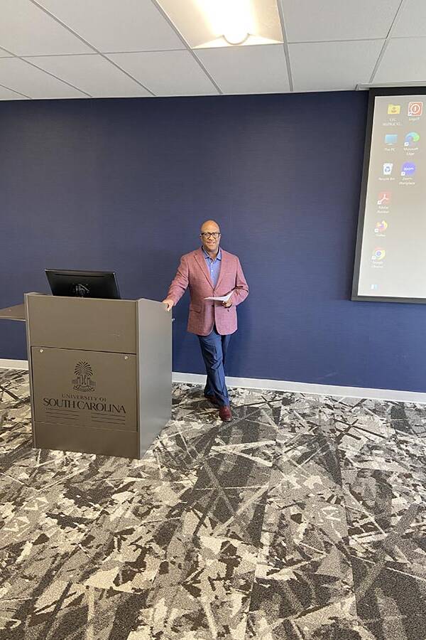 man in a mauve jacket and jeans leans one hand on a podium.