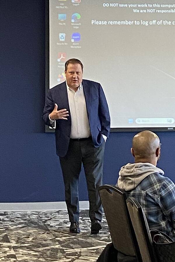 Man in a dark suit jacket standing near a drop down screen Another man in a hoodie is in the foreground.