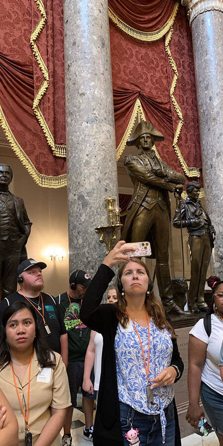 people standing in a room with large marble columns and bronze statues. Crimson and gold drapes hang in between columns.