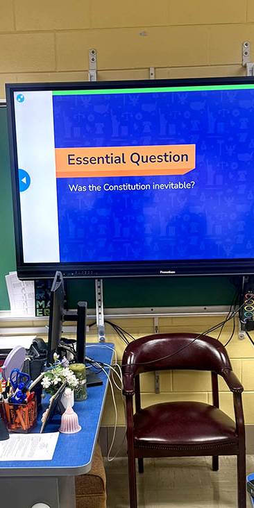 Teacher in orange stands to the right of a screen which says 