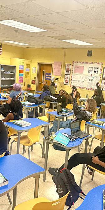 Teacher in orange leads class discussion in front of screen.