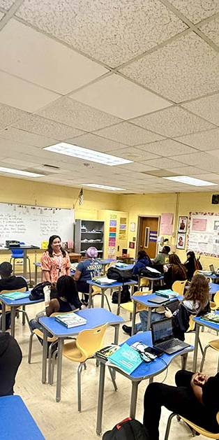 Teacher in orange leads class discussion in front of screen.