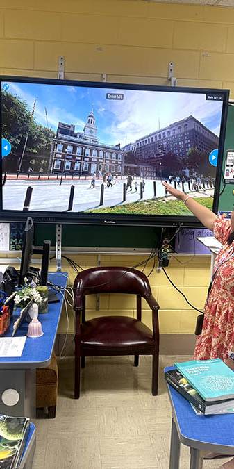 Teacher in orange leads class discussion in front of screen.
