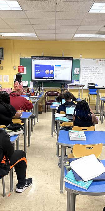 Teacher in orange leads class discussion in front of screen.