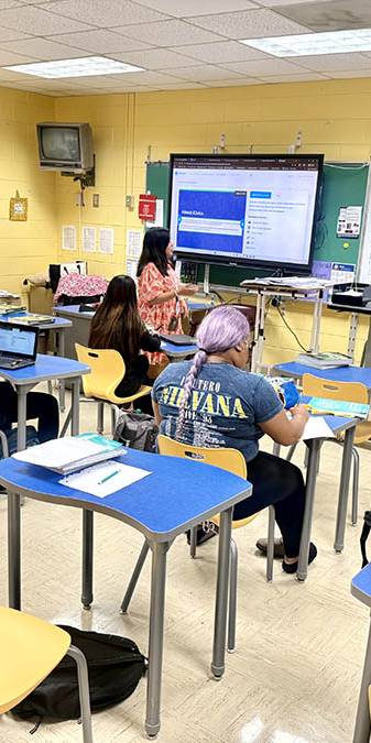 Teacher in orange leads class discussion in front of screen.