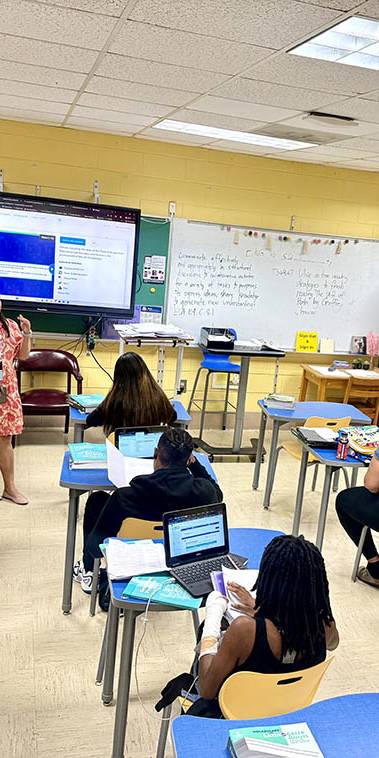 Teacher in orange leads class discussion in front of screen.