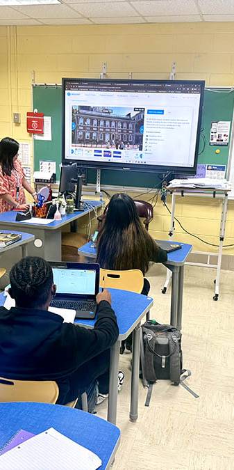 Teacher in orange leads class discussion in front of screen.